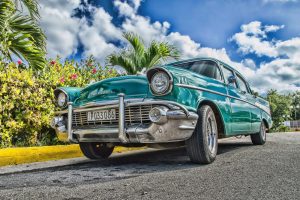 A beautifully restored classic car on a sunny road in Havana, Cuba, with lush foliage and a vibrant sky.